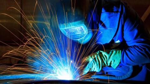 Close-up of welder at work fabricating metal in foreground, dressed in blue with sparks flying.